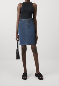 Vrouw met een zwarte mouwloze top, gespen denimrok, zwarte slippers, met een zwarte handtas tegen een effen lichte achtergrond.