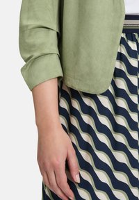 Veste en tissu texturé vert clair avec des manches retroussées, assortie à une jupe bleu marine ornée de bandes ondulées crème et olive.