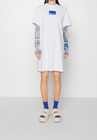 Weißes T-Shirt-Kleid mit blauem Logo, über einem langärmeligen, tie-dye Shirt getragen. Beigefarbene, flauschige Sandalen, blaue Socken. Einfaches und lässiges Design.