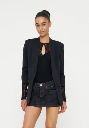 Kvinde med mørkt hår sat op i en knold iført en sort blazer, sort blonde top og mørk denim miniskørt, stående mod en ensfarvet lys baggrund.
