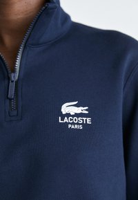 Mornarica dukserica na patent zatvarač od glatkog materijala sa bijelim Lacoste logom i natpisom "PARIS" na prednjem dijelu.