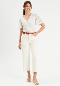 Blouse crème ample avec des détails en dentelle, associée à un pantalon large blanc taille haute et des sandales à talons dorées. Ceinture brune incluse.