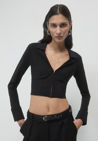 Cropped top nero con colletto, maniche lunghe e zip frontale. Presenta cuciture sartoriali e un tessuto liscio e testurizzato.