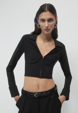 Cropped top nero con colletto, maniche lunghe e zip frontale. Presenta cuciture sartoriali e un tessuto liscio e testurizzato.