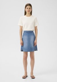 Jupe en denim en forme de A avec un lavage bleu, dotée d'une taille haute et de détails d'ourlet incurvé, associée à un t-shirt crème à manches courtes.