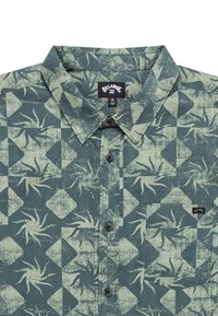 Chemise boutonnée à manches courtes de couleur vert foncé avec un motif abstrait de feuilles et de diamants en vert clair. Présente un col et des boutons noirs.