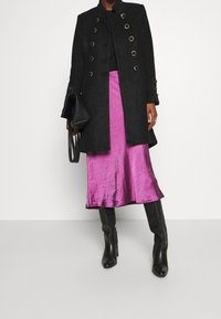 Abrigo negro entallado con acentos de botones dorados, combinado con una falda fucsia brillante. La modelo lleva botas de tacón negro hasta la rodilla y sostiene un bolso negro.