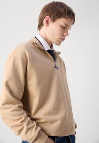 Felpa beige con zip, dal finish testurizzato, caratterizzata da un colletto alto e un piccolo logo blu, abbinata a una camicia bianca sotto.