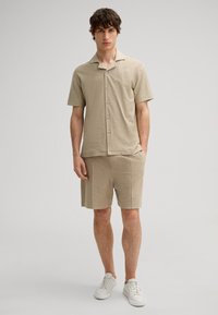 Beige kurzärmliges Button-up-Hemd mit strukturiertem Muster, kombiniert mit passenden Shorts, beide im lässigen Schnitt. Getragen mit weißen Sneakers.
