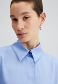 Jasnoniebieska koszula na guziki z wywiniętym kołnierzykiem, charakteryzująca się gładką teksturą i starannym przeszyciem. Modelka nosi srebrny kolczyk w kształcie kółka.