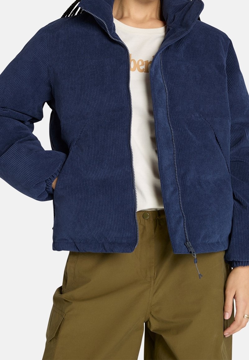 Personne portant une veste en velours côtelé bleu marine à fermeture éclair, un t-shirt blanc graphique et un pantalon cargo vert olive, les mains dans les poches de la veste.