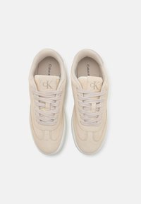 Beige semskinn joggesko med lysegrå snørebånd, med en glatt lærsåle og preget logo på tungen. Rundt tådesign.