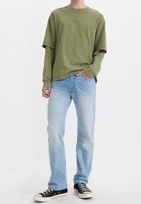 Camiseta de manga larga verde oliva con un ajuste relajado, combinada con jeans de pierna recta en azul claro y zapatillas negras bajas. Detalle de cadena plateada.