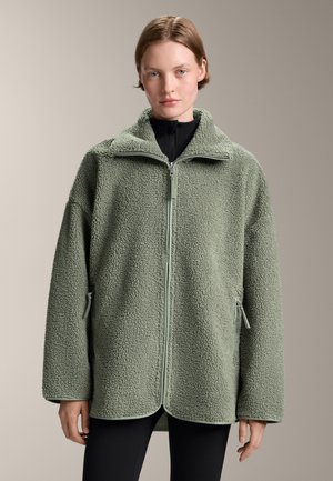 Groene fleecejack met een ritssluiting aan de voorkant, hoge kraag en zijzakken met rits. Zacht, gestructureerd materiaal en een losse pasvorm.