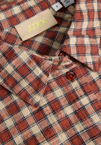 Rdeča in bež karo srajca z ovratnikom, ki ima majhne rjave gumbe in rumeno oznako z napisom "JJXX." Narejena iz teksturirane tkanine.
