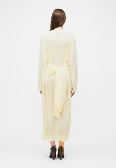 Γυναίκα με μακριά καστανά μαλλιά που φορεί εκρού μακρύ φόρεμα-πουλόβερ με δεμένη λεπτομέρεια κόμπο στο πίσω μέρος και σκούρα καφέ παπούτσια με τακούνι.