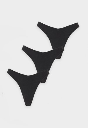 Femme debout pieds nus portant un caraco blanc bordé de dentelle et un dessous style bikini noir sur un fond gris uni.