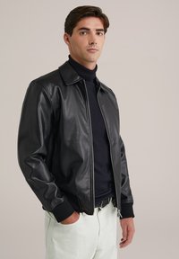 Chaqueta de cuero negra con cierre de cremallera y cuello puntiagudo, combinada con un suéter negro de cuello alto y pantalones de color claro. Textura suave.