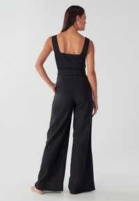 Žena v bezramenném černém overal s širokými nohavicemi, stojící s jednou rukou v kapse, čelící od záznamu proti jednobarevnému pozadí.