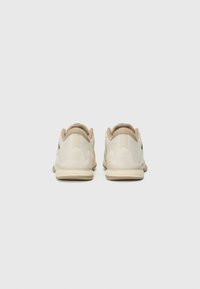 Baskets de sport beige avec une tige en mesh texturé, un col rembourré et une semelle extérieure en caoutchouc souple. Présente des accents de logo subtils au niveau du talon.