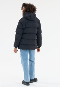 Person mit lockigem Haar, der eine schwarze Steppjacke mit Kapuze, hellblaue Jeans und schwarze Stiefel trägt, steht mit dem Rücken zu einer weißen Hintergrund.
