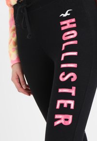 Zbliżenie na czarne spodnie z różowym napisem i logo "Hollister" na udzie oraz ręką spoczywającą obok nogi ubraną w wielokolorowy rękaw.