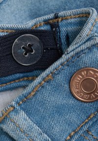Denim tkanina u svijetloplavoj boji, s tamnoplavim elastičnim pojasom, prozirnim plastičnim dugmetom i metalnim dugmetom boje bakra s utisnutim tekstom.