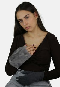 Szare rękawiczki z futrzanym sztucznym futerkiem o miękkiej fakturze po jednej stronie i gładkim, ciemnym materiale po drugiej, noszone z brązową bluzką i szarymi dżinsami.