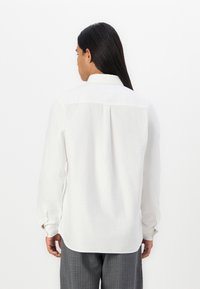 White long-sleeve shirt with a collar, featuring a yoke and a central back pleat. The fabric appears lightweight with a smooth texture.