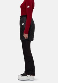 Black insulated skirt with quilted pattern, paired with a red long-sleeve top. Black pants and white ski boots with black accents complete the look.