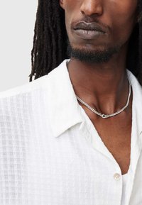 Camicia bianca strutturata con colletto corto, dotata di chiusura con bottoni. Il modello indossa una collana in argento che valorizza il scollo.