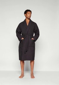 Black waffle-textured bathrobe with long sleeves and a tied waist. Features two front pockets and a shawl collar, resting above bare feet.