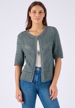 Vrouw draagt een petrolkleurige gebreide cardigan met korte mouwen en een enkele bovenste knoop, gecombineerd met blauwe spijkerbroek en een wit topje.