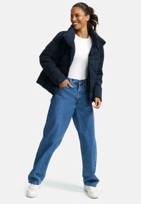 Marinblå quiltad pufferjacka med hög krage, bärs över en vit skjorta, i kombination med lösa blå jeans och vita sneakers.