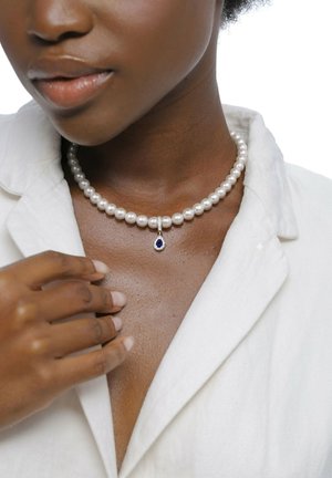 Mørkhudet kvinde iført en hvid blazer og en perlehalskæde med en blå dråbevedhæng, der forsigtigt rører ved halskæden.