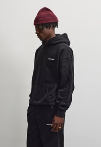 Junger Mann, der einen schwarzen Pegador-Hoodie und eine Jogginghose sowie eine dunkelrote gestrickte Mütze trägt, steht seitlich vor einem schlichten hellen Hintergrund.