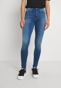 Jeans ajustados de mezclilla en color azul, con una cintura alta, bolsillos frontales y un acabado desgastado con una textura suave. Combinados con zapatillas negras.