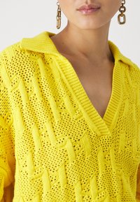 Gul stickad tröja med V-hals, öppen vävstruktur och texturerad design. Krage med detaljer ger visuell intress.