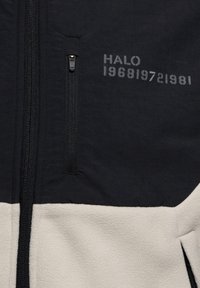 Fleecejacka med en svart övre del och en krämfärgad nedre del. Inkluderar fickor med dragkedja och tryckt text: "HALO 196819721981."