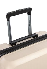Valigia rigida beige con superficie testurizzata, zip nera e manico retrattile argento etichettato "SURI FREY."