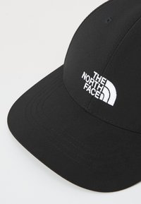 Svart baseballcaps laget av stoff med buet skjerm. Har hvit brodert logo "THE NORTH FACE" på forsiden. Glatt tekstur.