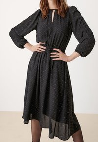 Žena v černých midi šatech se zlatými puntíky, dlouhými nabíranými rukávy, výstřihem ve tvaru klíčové dírky a asymetrickým lemem, s rukama na bocích.