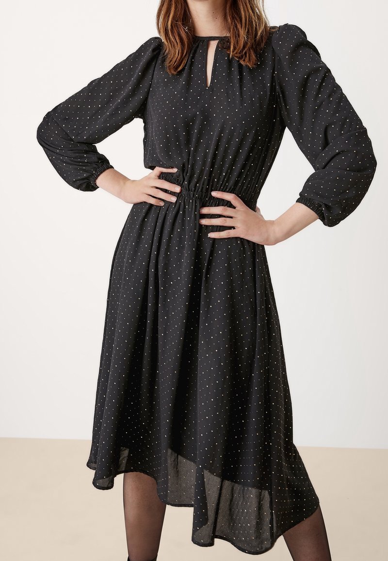 Žena v černých midi šatech se zlatými puntíky, dlouhými nabíranými rukávy, výstřihem ve tvaru klíčové dírky a asymetrickým lemem, s rukama na bocích.