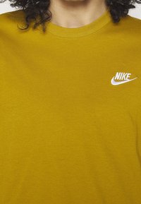 Hořčičkově žluté bavlněné tričko s kulatým výstřihem a malým bílým logem Nike na levé straně hrudi. Měkká textura a krátké rukávy.