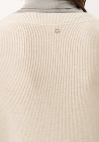 Lichtbeige gebreide trui met textuur en een geribbelde structuur, met een zilveren ronde accent aan de achterkant van de nek. Zacht, ademend materiaal.