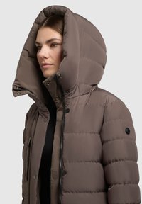 Braune Steppjacke mit großer Kapuze. Verfügt über eine gesteppte Textur, Reißverschluss und Knopfdetails. Gestaltet mit hohem Kragen und seitlichen Taschen.