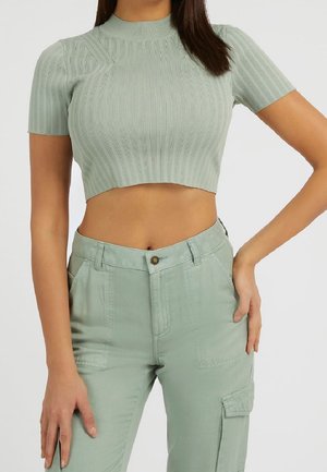 Vrouw draagt een lichtgroen geribd crop topje met korte mouwen en bijpassende cargobroek met hoge taille, met één hand op haar heup.