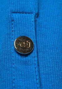 Blå ribbad tyg med en framträdande rund guldknapp med en design av en symaskin. Texturerad yta med synliga sömnadsdetaljer.