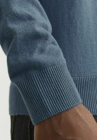 Hellblauer gestrickter Pullover mit gerippten Bündchen und glatter Textur, teilweise auf einem dunklen Arm vor einem neutralen Hintergrund zu sehen.