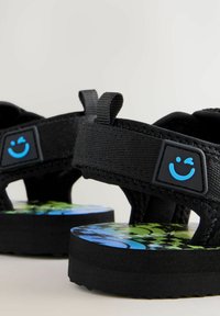 Svarta sandaler med texturerade tygremmar, justerbara stängningar och en blå leende ikon. Färgglad tryckt fotbädd har gröna och blå mönster.